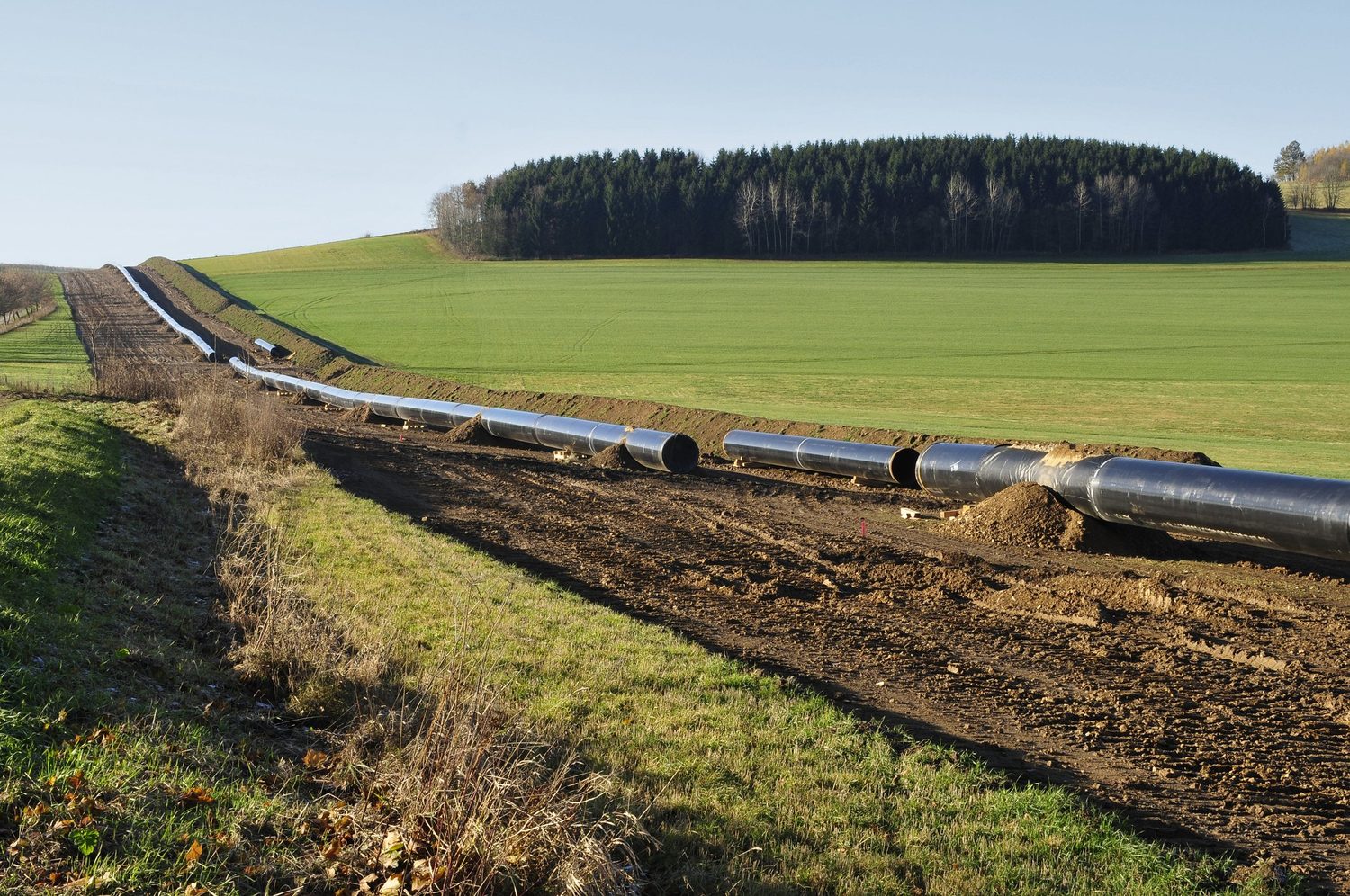 A pipeline under construction stretching across rural farmland and into the treeline beyond — resources infrastructure crossing the country whose consent it needs.